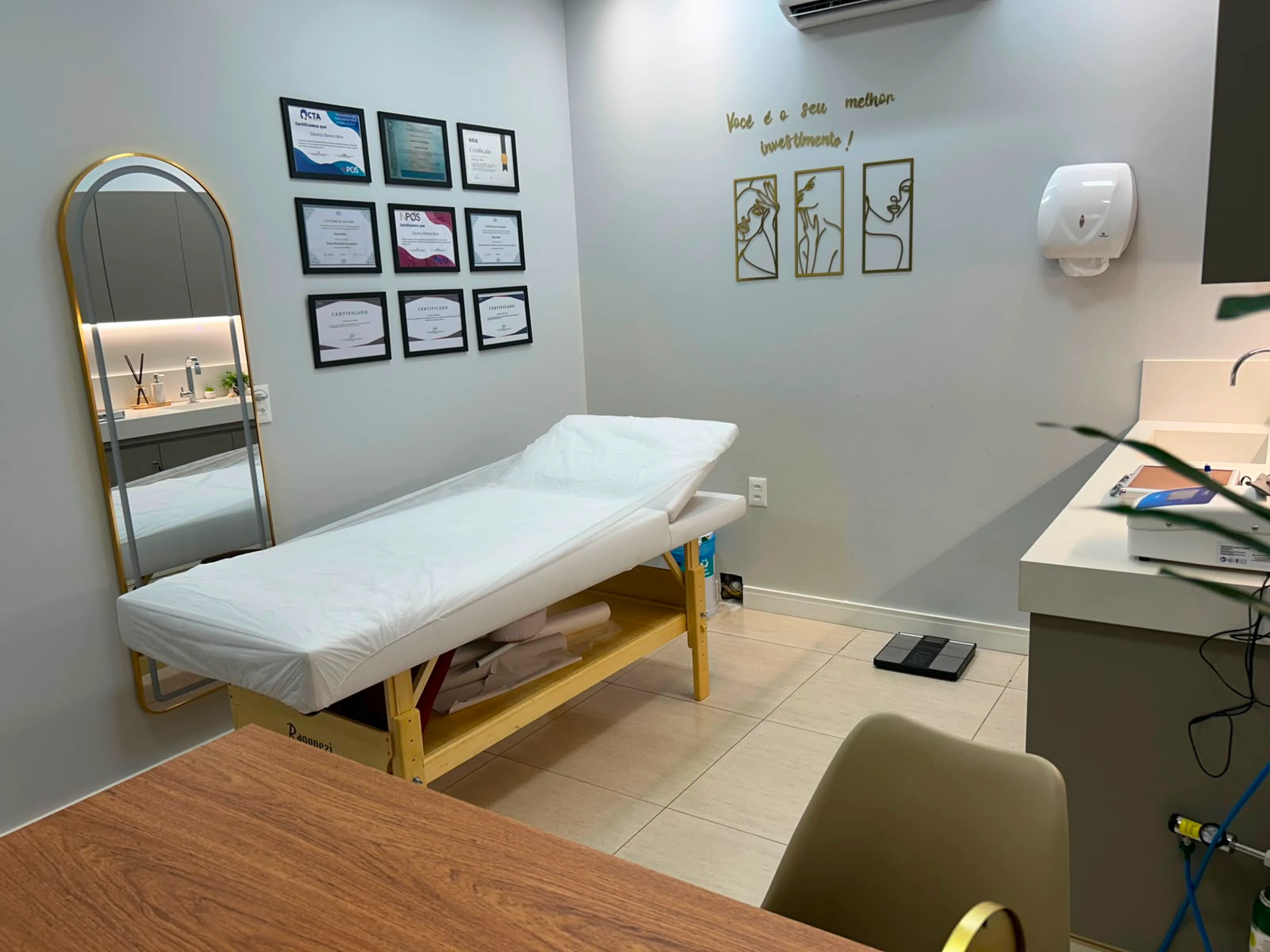 Sala de estética com maca, espelho dourado e diplomas na parede